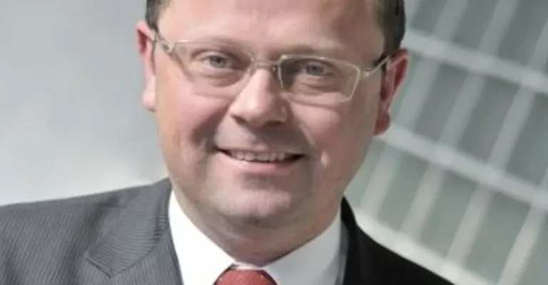 Mężczyzna w garniturze i czerwonym krawacie, uśmiechnięty, na tle szklanej, nowoczesnej fasady budynku.