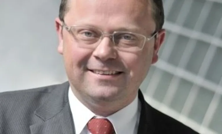 Mężczyzna w garniturze i czerwonym krawacie, uśmiechnięty, na tle szklanej, nowoczesnej fasady budynku.