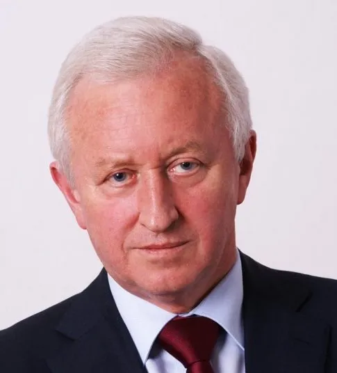 Głowa i ramiona starszego mężczyzny o siwych włosach, w garniturze i czerwonym krawacie, na jasnym tle.