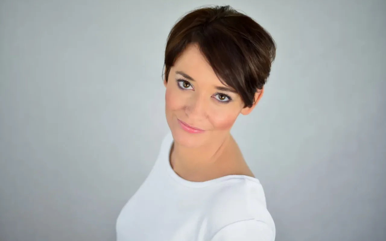 Portret kobiety z krótkimi ciemnymi włosami, uśmiechniętej, w białej bluzce, na neutralnym, jasnym tle.