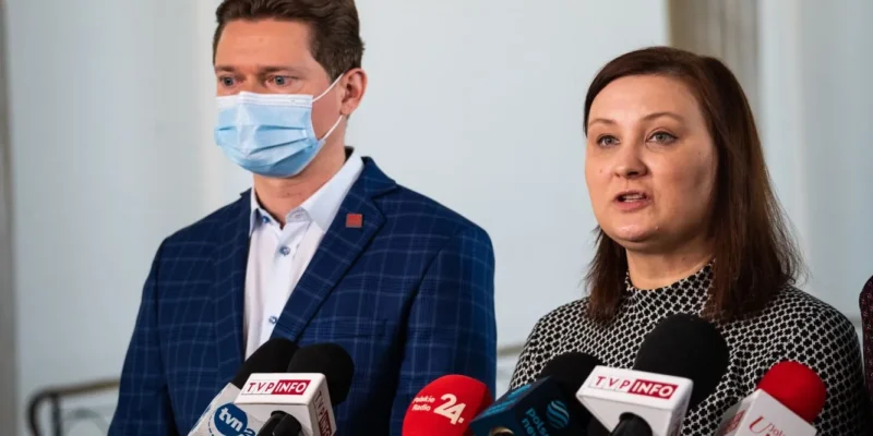 Dwóch mówców na konferencji prasowej stojących przy mikrofonach; mężczyzna w maseczce ochronnej i granatowej kratkowanej marynarce, kobieta po jego prawej stronie mówi.