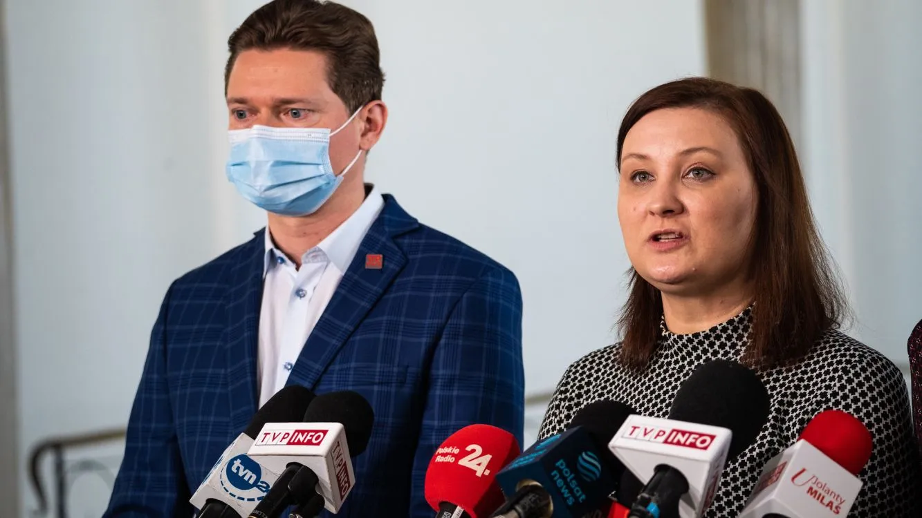 Dwóch mówców na konferencji prasowej stojących przy mikrofonach; mężczyzna w maseczce ochronnej i granatowej kratkowanej marynarce, kobieta po jego prawej stronie mówi.