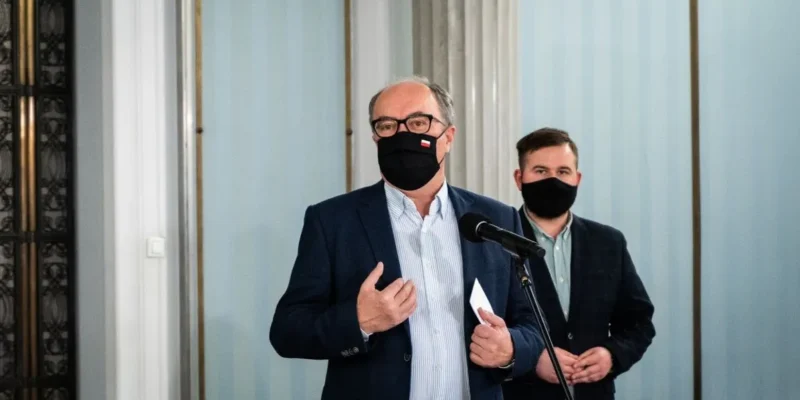 Dwóch mężczyzn w garniturach i maseczkach stoi przy mikrofonie, jeden gestykuluje i trzyma kartkę.