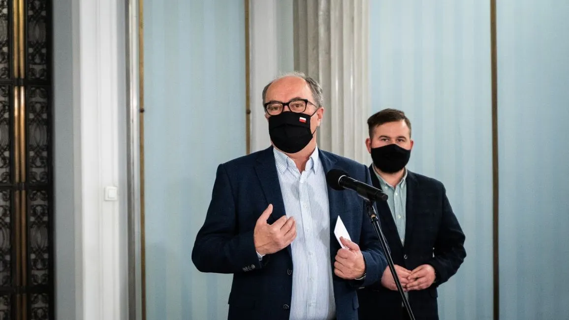 Dwóch mężczyzn w garniturach i maseczkach stoi przy mikrofonie, jeden gestykuluje i trzyma kartkę.