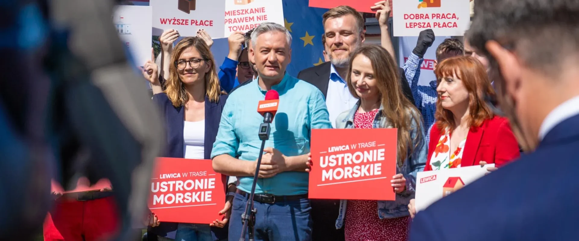 Grupa osób na konferencji prasowej Lewicy trzyma plakaty z hasłami wyborczymi, stojąc wokół mikrofonu na tle banerów i flag UE.