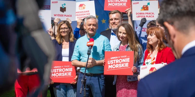 Grupa osób na konferencji prasowej Lewicy trzyma plakaty z hasłami wyborczymi, stojąc wokół mikrofonu na tle banerów i flag UE.
