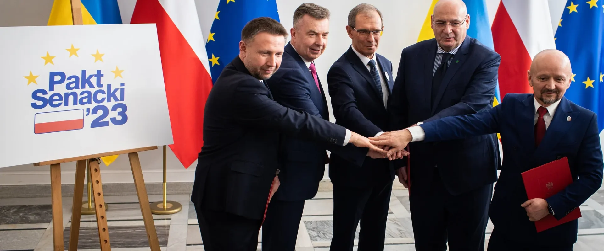Grupa mężczyzn w garniturach łączy dłonie w geście jedności podczas podpisywania Paktu Senackiego ’23, stojąc obok planszy z napisem i flag w tle.