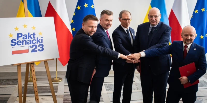 Grupa mężczyzn w garniturach łączy dłonie w geście jedności podczas podpisywania Paktu Senackiego ’23, stojąc obok planszy z napisem i flag w tle.
