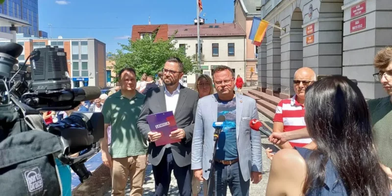 Grupa osób udziela konferencji prasowej przed urzędem miasta Pacanów, a dziennikarze z kamerą i mikrofonami relacjonują wydarzenie.