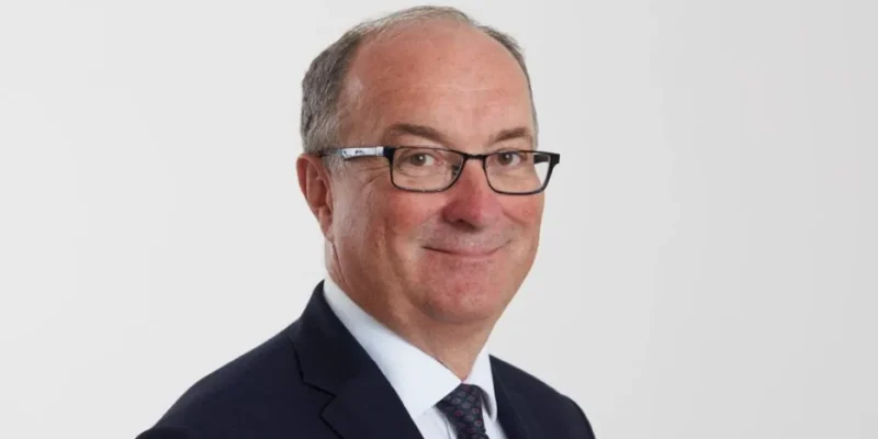 Portret mężczyzny w garniturze i okularach na jasnym tle, z delikatnym uśmiechem.