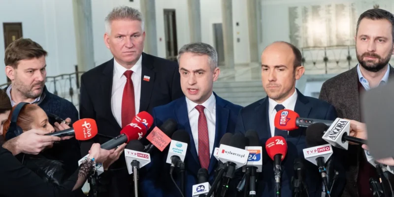 Na zdjęciu widoczna jest grupa polityków podczas konferencji prasowej, otoczona mediami w tle budynku.