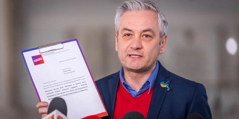 Mężczyzna w marynarce i czerwonym swetrze trzyma dokument w ręku, przemawiając do dziennikarzy w otoczeniu mikrofonów.