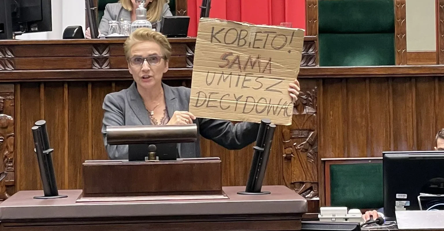 Na zdjęciu osoba trzymająca kartonowy plakat z napisem „KOBIETO! SAMA UMIESZ ZDECYDOWAĆ” przemawia w parlamencie.