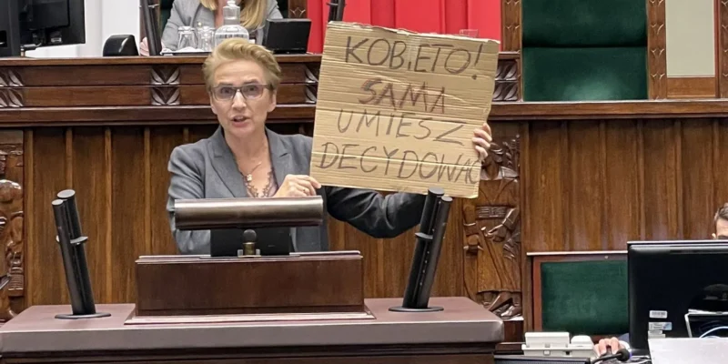 Na zdjęciu osoba trzymająca kartonowy plakat z napisem „KOBIETO! SAMA UMIESZ ZDECYDOWAĆ” przemawia w parlamencie.