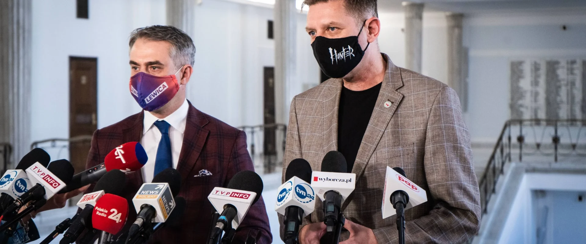 Dwóch mężczyzn w garniturach udziela konferencji prasowej w budynku z kolumnami, stojąc przed licznymi mikrofonami; po lewej maseczce z napisem „LEWICA”, po prawej maseczce z napisem „HUNTER”.