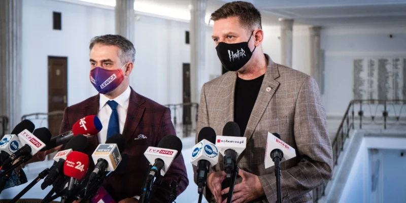 Dwóch mężczyzn w garniturach udziela konferencji prasowej w budynku z kolumnami, stojąc przed licznymi mikrofonami; po lewej maseczce z napisem „LEWICA”, po prawej maseczce z napisem „HUNTER”.