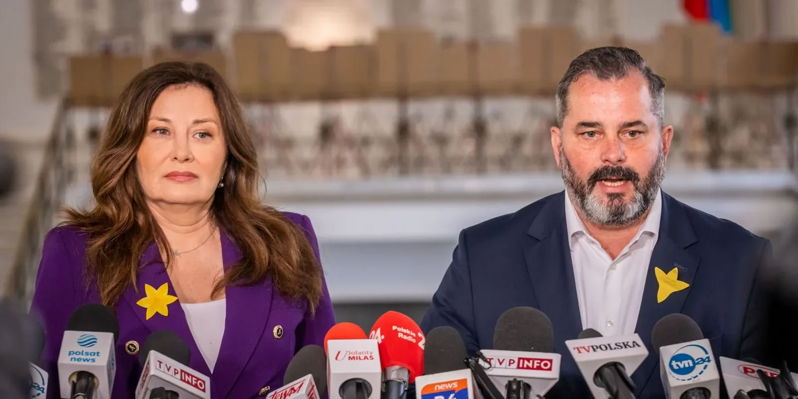 Na zdjęciu przedstawiono dwoje polityków podczas konferencji prasowej, z mikrofonami i kamerami skierowanymi na nich.