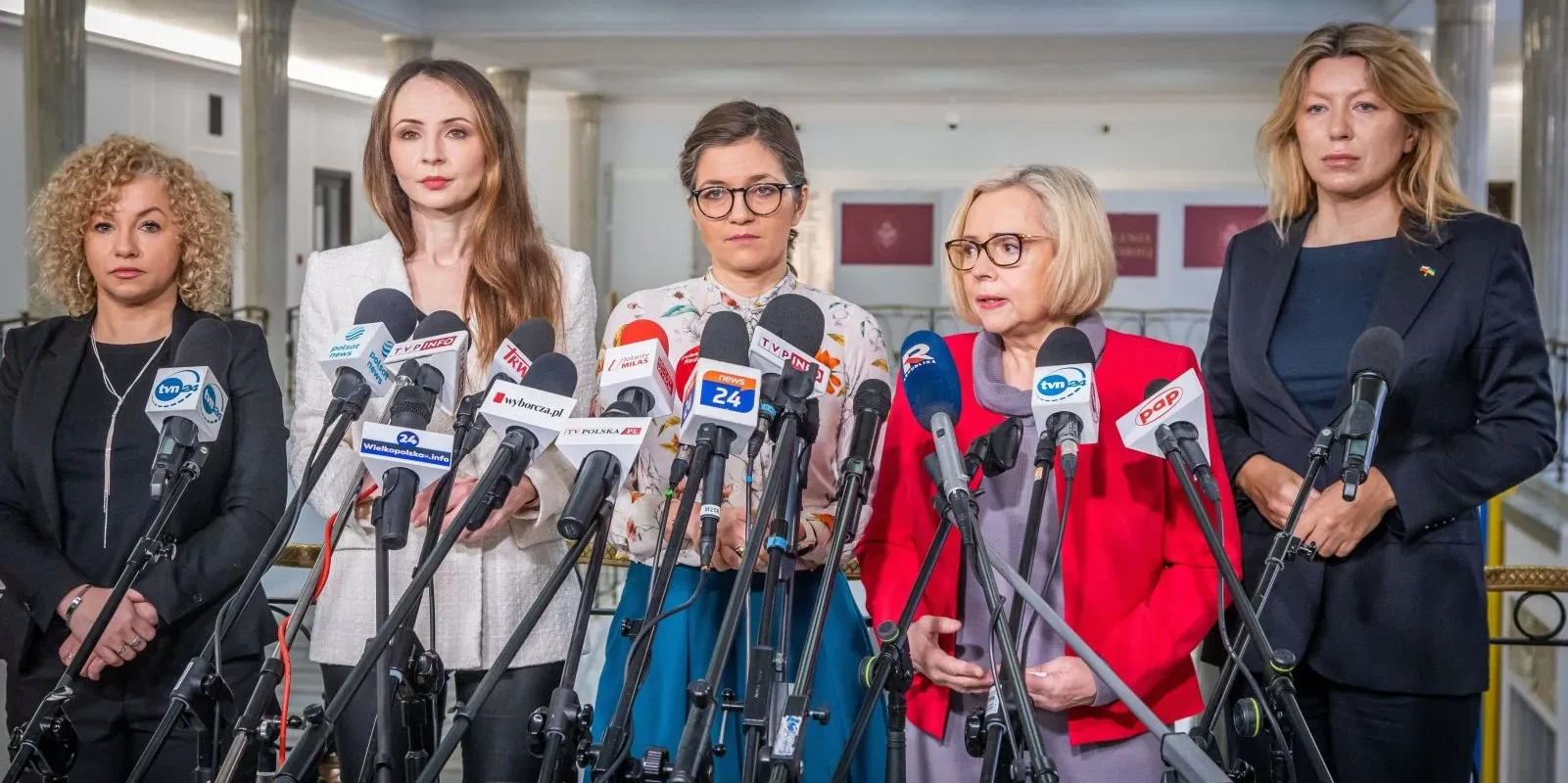 Na zdjęciu widoczne jest kilka kobiet stojących za mikrofonami podczas konferencji prasowej w budynku instytucji państwowej.