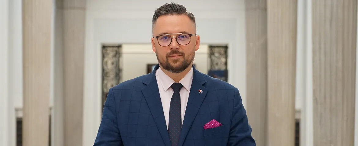 Mężczyzna w granatowym garniturze i okularach stoi w korytarzu z kolumnami.