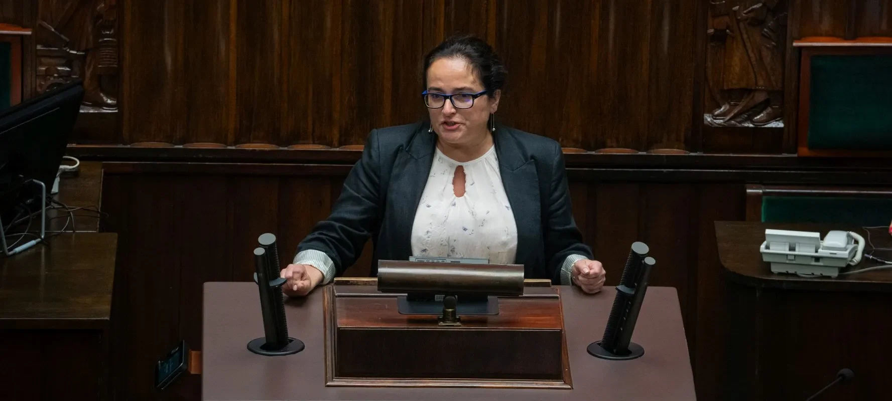 Kobieta w czarnym garniturze przemawia z mównicy w sali posiedzeń, otoczona drewnianymi panelami i mikrofonami.