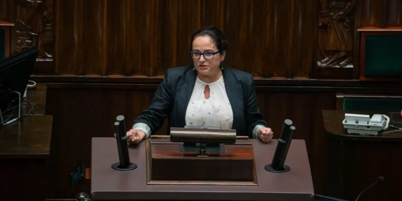 Kobieta w czarnym garniturze przemawia z mównicy w sali posiedzeń, otoczona drewnianymi panelami i mikrofonami.