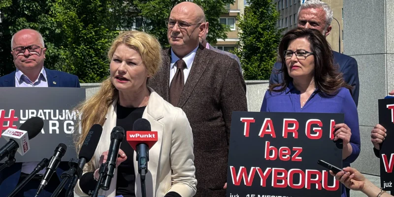 Grupa osób podczas konferencji prasowej trzyma plakaty z napisem „TARGI bez WYBORU” i apeluje do rządu o konkretne daty otwarcia branży targowej oraz wsparcie finansowe.