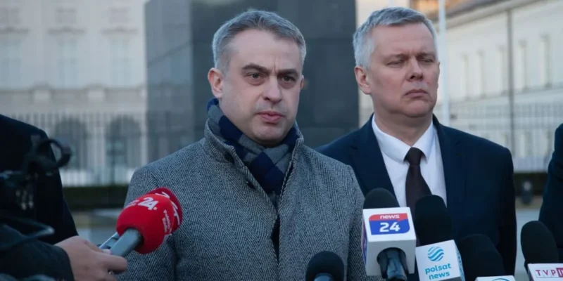 Na zdjęciu widać dwóch mężczyzn, którzy stoją przed mikrofonami i udzielają wywiadu, a w tle znajduje się budynek z jasnymi elewacjami.