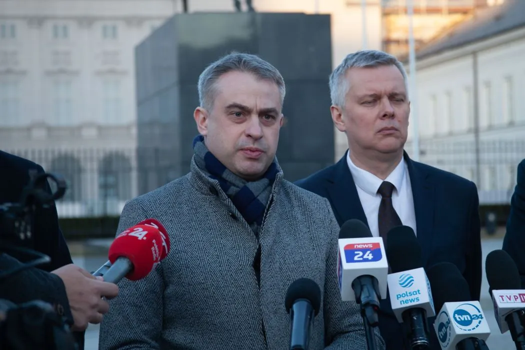 Na zdjęciu widać dwóch mężczyzn, którzy stoją przed mikrofonami i udzielają wywiadu, a w tle znajduje się budynek z jasnymi elewacjami.