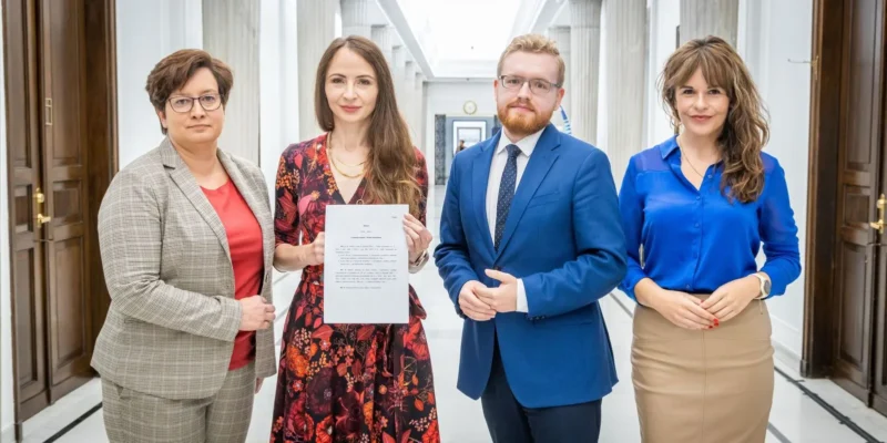 Na zdjęciu cztery osoby stoją w korytarzu budynku, trzymając dokument, w formalnych ubraniach.