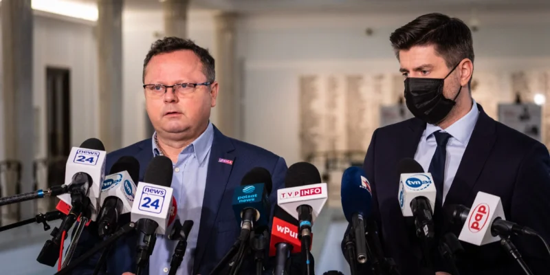 Dwóch mężczyzn w garniturach podczas konferencji prasowej, otoczonych licznymi mikrofonami z logotypami mediów; jeden bez maseczki, drugi w czarnej maseczce.