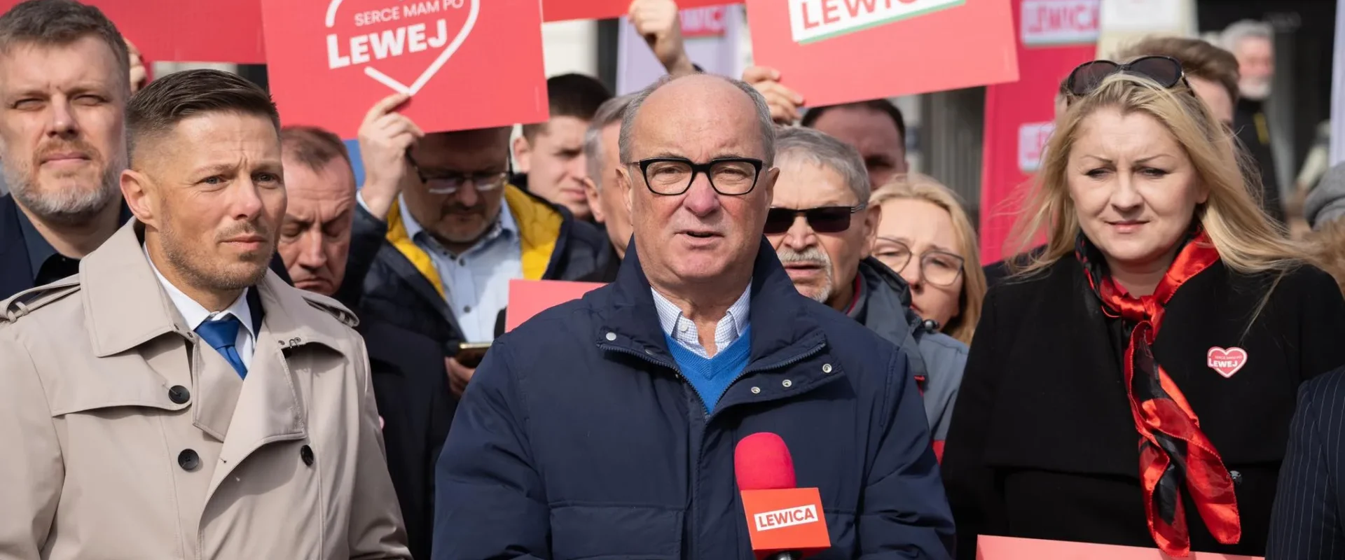 Na pierwszym planie mężczyzna w okularach mówi do mikrofonu z logiem Lewica, otoczony grupą osób trzymających czerwone plakaty z napisem LEWICA.