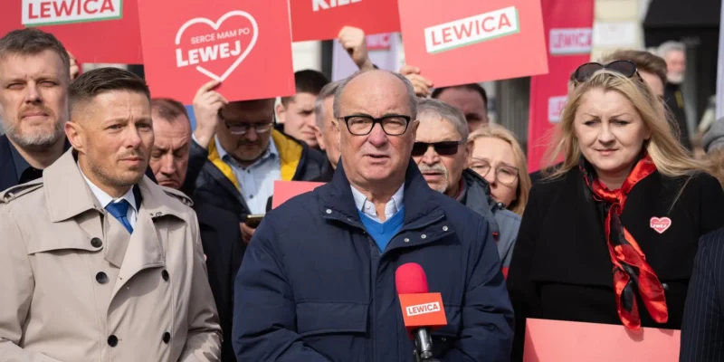 Na pierwszym planie mężczyzna w okularach mówi do mikrofonu z logiem Lewica, otoczony grupą osób trzymających czerwone plakaty z napisem LEWICA.