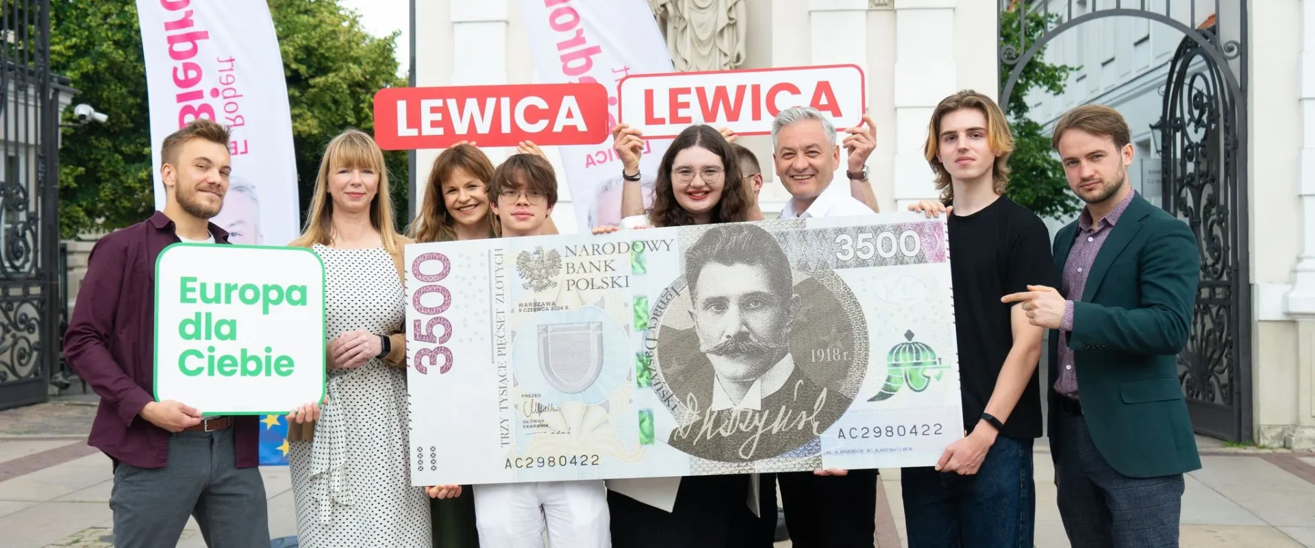 Grupa osób stoi przed bramą, trzymając duży plakat w kształcie banknotu z napisem 3500 zł oraz banery Lewica i znak „Europa dla Ciebie”.