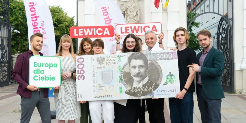 Grupa osób stoi przed bramą, trzymając duży plakat w kształcie banknotu z napisem 3500 zł oraz banery Lewica i znak „Europa dla Ciebie”.