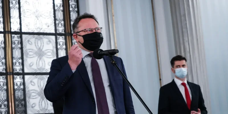 Dwóch mężczyzn w garniturach i maseczkach stoi przy mikrofonie podczas konferencji; pierwszy przemawia, drugi stoi z boku w tle przy kolumnach i ozdobnych drzwiach.