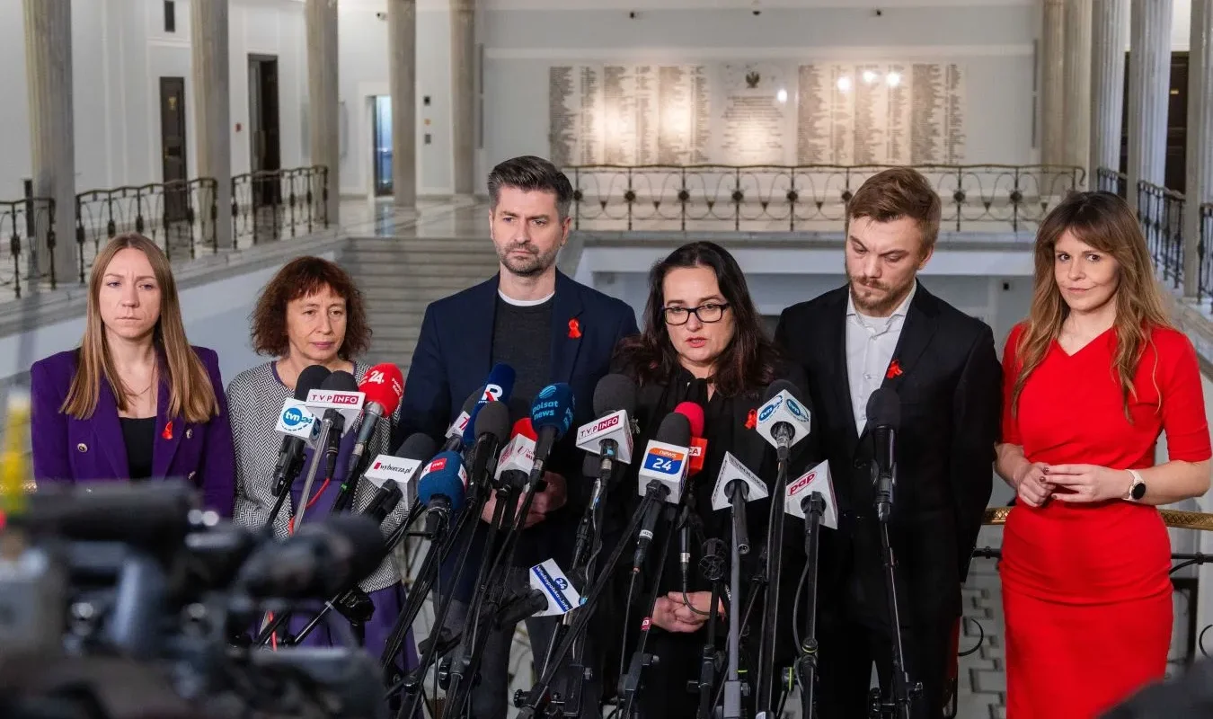 Grupa osób z czerwonymi wstążkami na piersiach stoi podczas konferencji prasowej z licznymi mikrofonami w holu, omawiając rekord zakażeń HIV w Polsce i potrzebę profilaktyki.