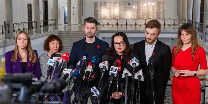 Grupa osób z czerwonymi wstążkami na piersiach stoi podczas konferencji prasowej z licznymi mikrofonami w holu, omawiając rekord zakażeń HIV w Polsce i potrzebę profilaktyki.