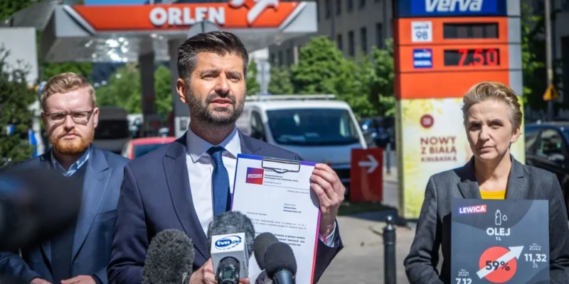 Konferencja prasowa na tle stacji paliw Orlen; mężczyzna w garniturze trzyma dokumenty i rozmawia z mediami, otoczony innymi osobami.