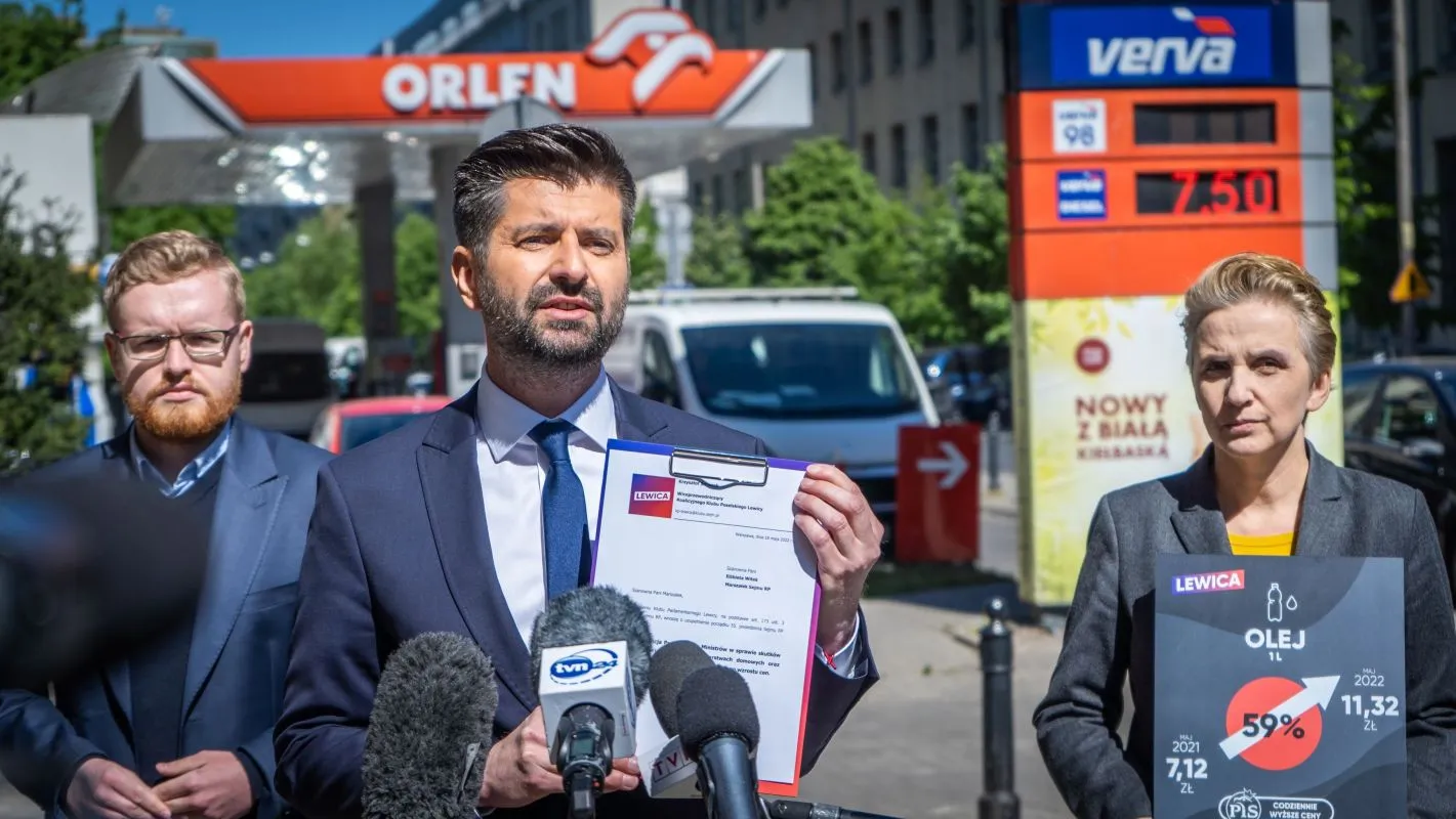 Konferencja prasowa na tle stacji paliw Orlen; mężczyzna w garniturze trzyma dokumenty i rozmawia z mediami, otoczony innymi osobami.