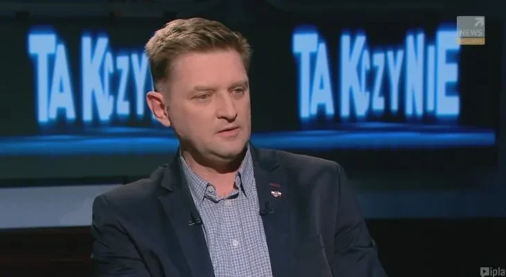 Mężczyzna w marynarce i koszuli siedzi w studiu telewizyjnym podczas publicystycznej rozmowy, a w tle widoczny jest duży podświetlony napis.