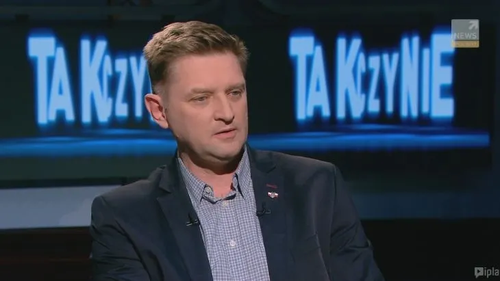 Mężczyzna w marynarce i koszuli siedzi w studiu telewizyjnym podczas publicystycznej rozmowy, a w tle widoczny jest duży podświetlony napis.