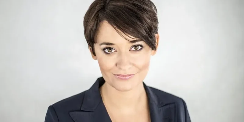 Portret kobiety o krótkich brązowych włosach, uśmiechniętej, w ciemnym żakiecie na jasnym, neutralnym tle.