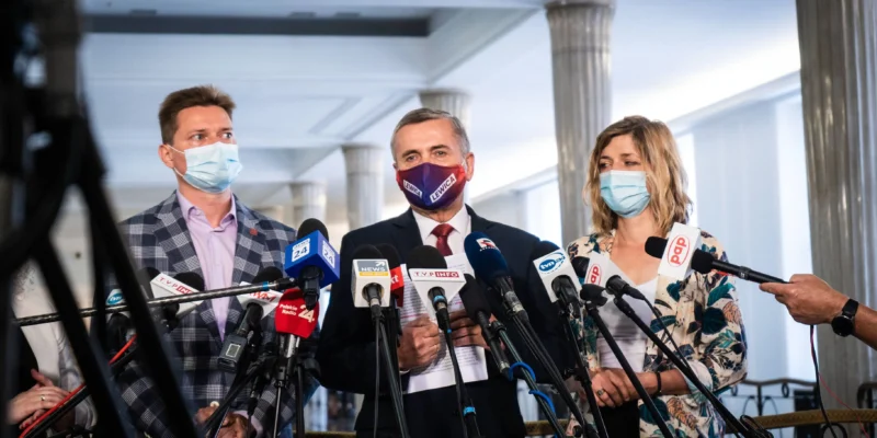 Grupa osób w maseczkach na konferencji prasowej stojąca za licznymi mikrofonami, omawia emerytury stażowe po 35 latach pracy kobiet i 40 latach pracy mężczyzn.