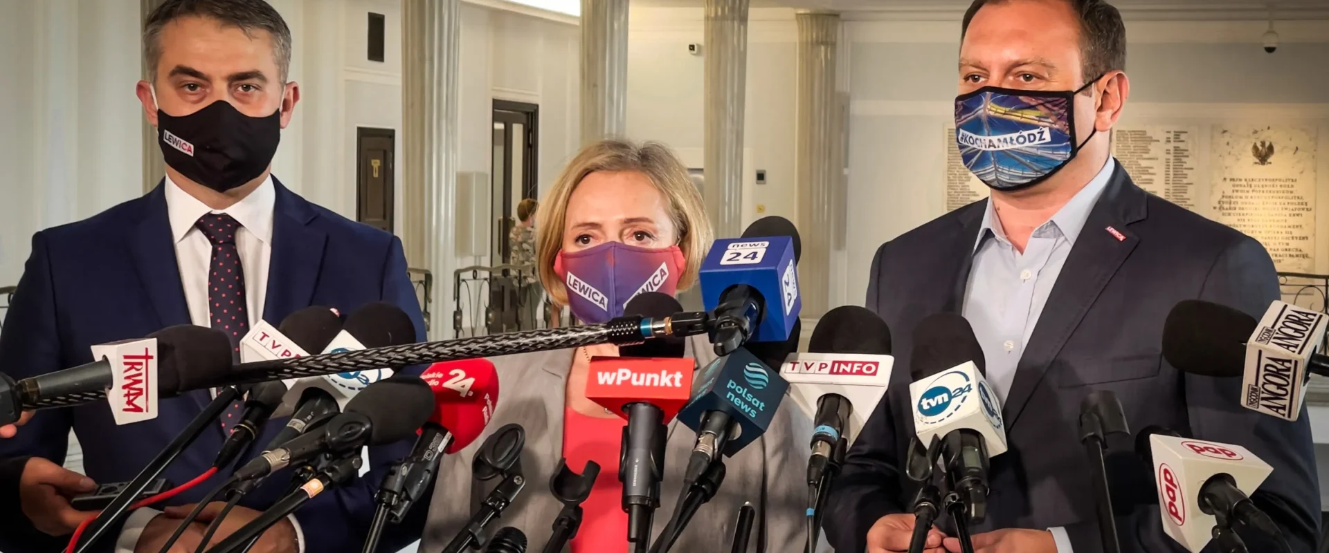 Trzech polityków w garniturach i maseczkach z napisem Lewica stoi przed licznymi mikrofonami podczas konferencji prasowej.