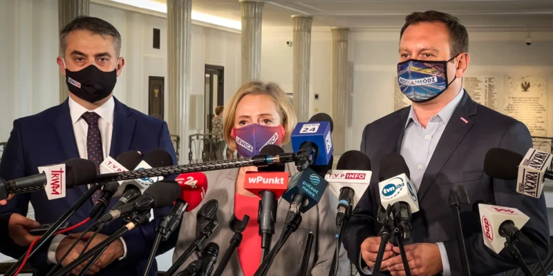 Trzech polityków w garniturach i maseczkach z napisem Lewica stoi przed licznymi mikrofonami podczas konferencji prasowej.