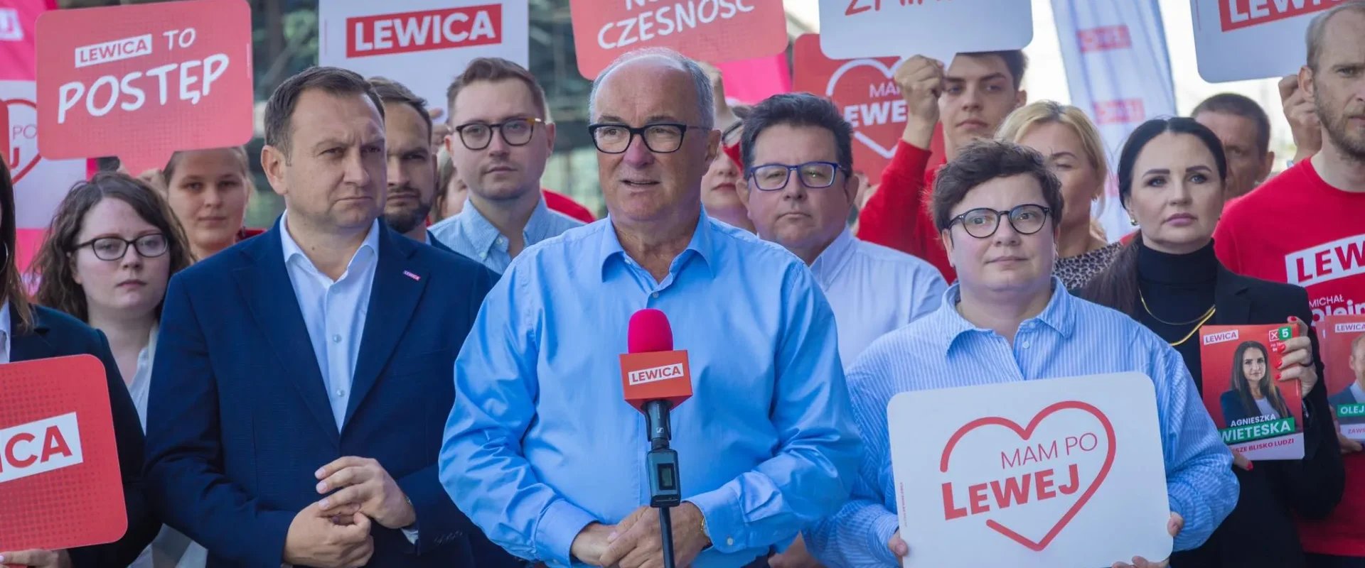 Grupa zwolenników Lewicy podczas plenerowej konferencji prasowej; na pierwszym planie mężczyzna w niebieskiej koszuli stoi przy mikrofonie, otoczony plakatami i banerami z hasłem Lewica.