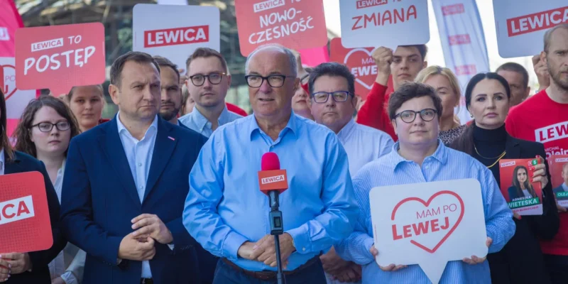 Grupa zwolenników Lewicy podczas plenerowej konferencji prasowej; na pierwszym planie mężczyzna w niebieskiej koszuli stoi przy mikrofonie, otoczony plakatami i banerami z hasłem Lewica.