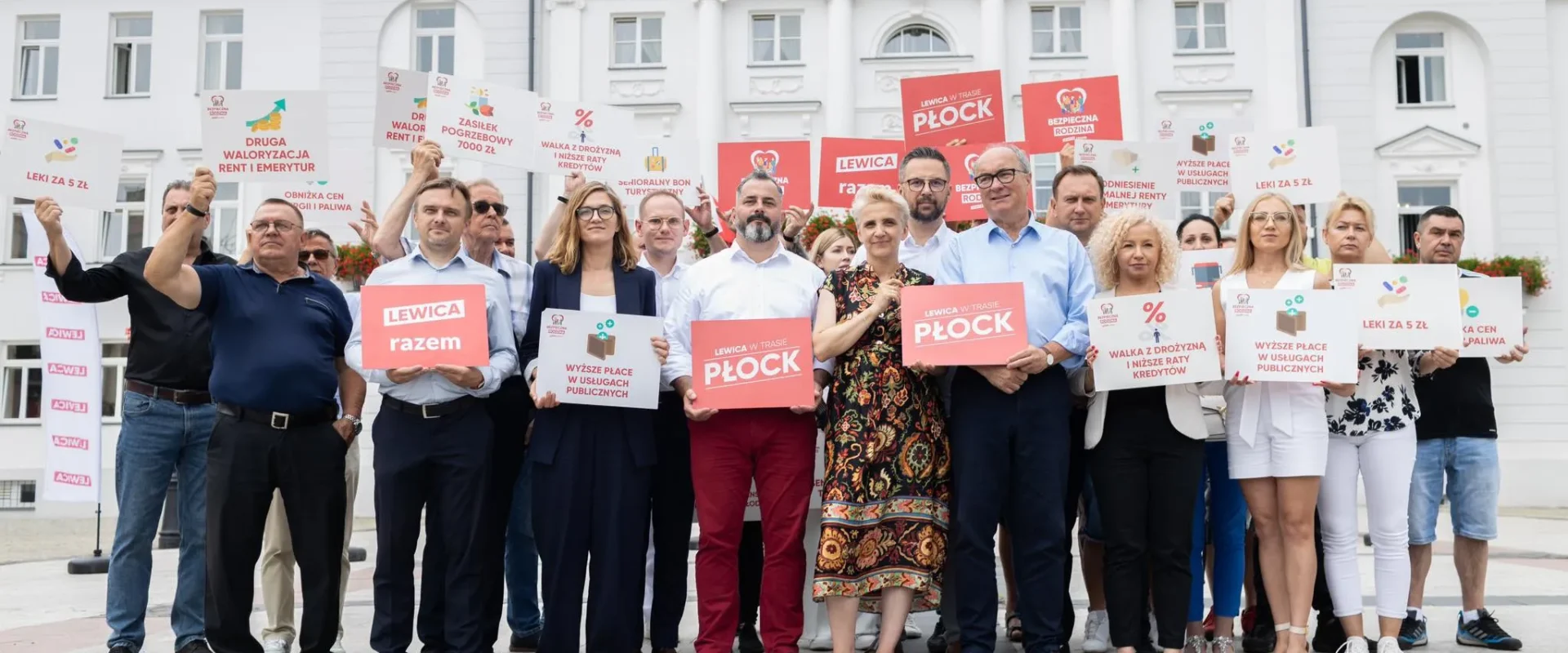 Grupa zwolenników Lewicy stoi na placu przed białym budynkiem, trzymając plakaty z hasłami o prawach pracowniczych i skróceniu czasu pracy.