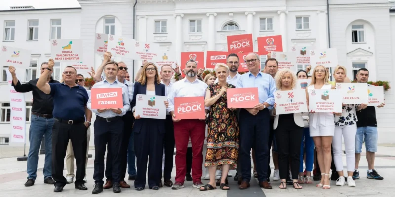 Grupa zwolenników Lewicy stoi na placu przed białym budynkiem, trzymając plakaty z hasłami o prawach pracowniczych i skróceniu czasu pracy.