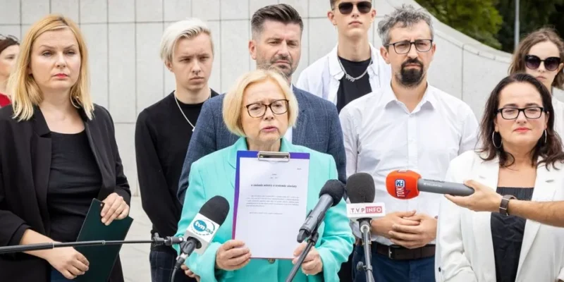 Na zdjęciu grupa osób stoi przed kamerami, z tą, która trzyma dokument, wyróżniającą się w centralnej części kadru.